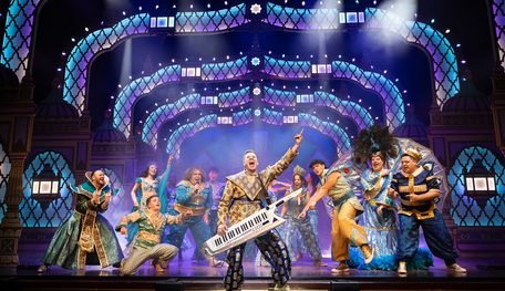 A person holding a keytar points to the sky and sings. Others behind point, shout and sing along, some holding microphones.