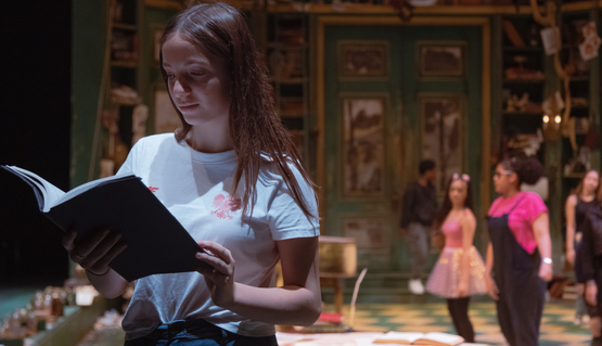 A young person reads from an open book whilst on the set of a theatre stage. A red logo reads: Christmas Challenge Big Give