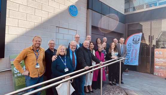 A group of people smile as they stand underneath a blue plaque, mounted to the concrete side of a theatre building.