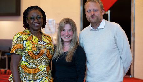 Bookey Oshin, Elizabeth Newman and Tom Bird are stood together in a theatre foyer. Behind is a large door to the theatre auditorium denoted by a red triangle