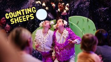 Text reads 'Counting Sheep'. Three actors on stage, performing in front of a crowd. Two actors are dressed as sheep and looking up. Another actor is above them holding white items above them.
