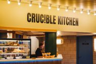 Crucible Kitchen lit up with bright celling spotlights. A kiosk-style cafe with an open counter display with cakes and sandwiches visible.