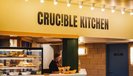 Crucible Kitchen lit up with bright celling spotlights. A kiosk-style cafe with an open counter display with cakes and sandwiches visible.