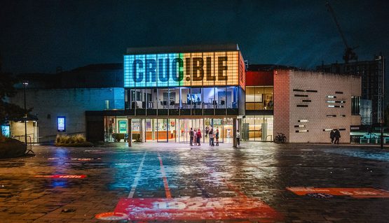The Crucible theatre is lit up at night in blue and orange light. On the floor in front, a brightly coloured race track leads up to the front of the theatre.