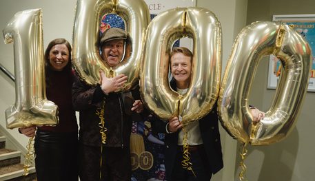 Three people smile with 1000 number balloons, two poking their heads through the 0s