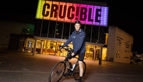 A person wearing riding clothing and a helmet poses on a bike outside the Crucible theatre. It is night-time and the theatre's sign is lit up in rainbow colours.