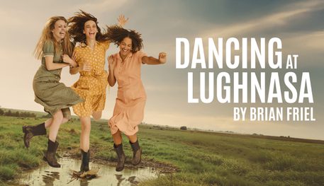 Three people with long hair and wearing pastel coloured dresses smile and laugh as they hold hands and jump into a puddle in the middle of a grey green field.