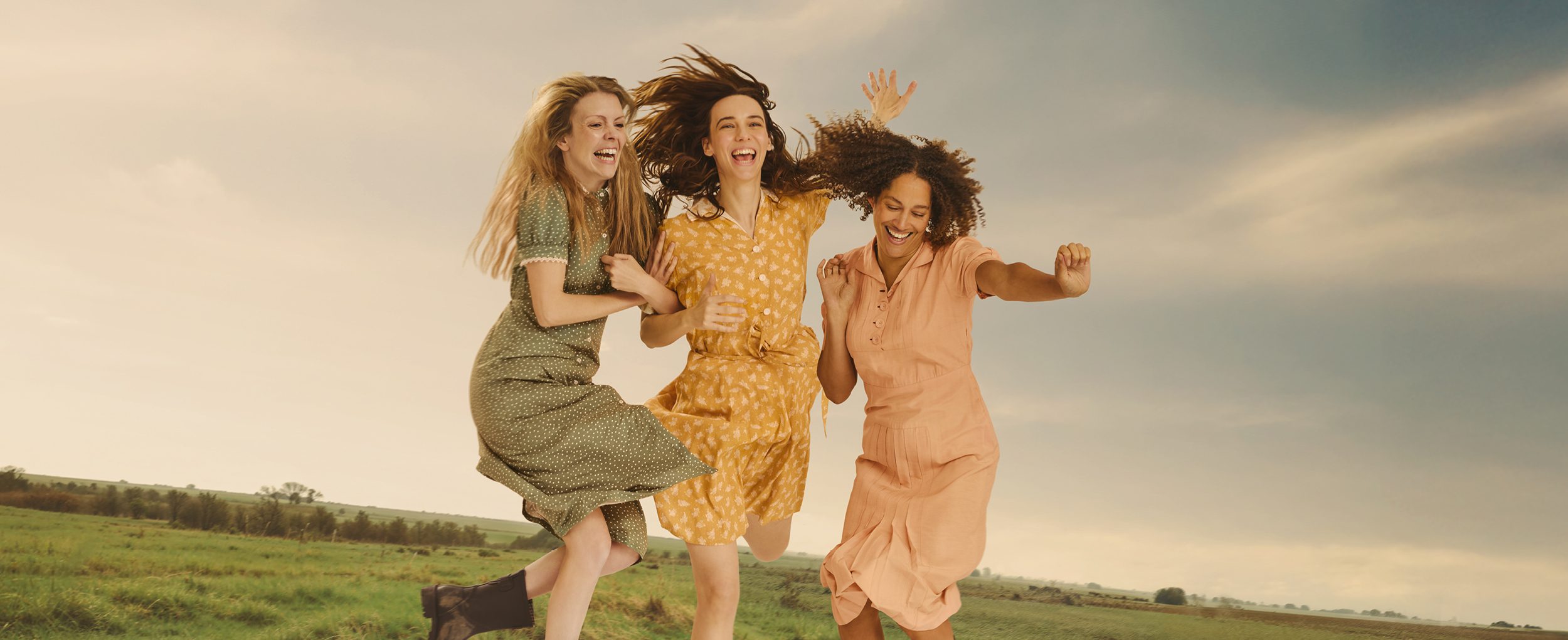 Three people with long hair and wearing pastel-coloured dresses smile and laugh as they hold hands and jump into a puddle in the middle of a grey green field.