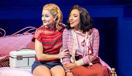Natasha Mould (Judy) and Grace Mouat (Betty) in Irving Berlin’s White Christmas. Photo by Johan Persson. Natasha and Grace sit side by side on a bed, holding each other’s arms as they listen.