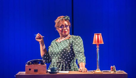 Sandra Marvin (Martha) in Irving Berlin’s White Christmas. Photo by Johan Persson. Sandra wears a polka dot dress and holds a pen in one hand and leans on a reception desk with the other. She smiles wryly.