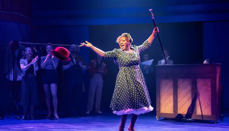 Sandra Marvin (Martha) in Irving Berlin’s White Christmas. Photo by Johan Persson. Sandra wears a polka dot dress and flings a red bowler hat across the room as she sings. In the other hand, she holds a cane and she is smiling triumphantly.