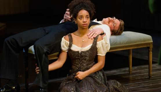 Siena Kelly (Nora) and Tom Glenister (Torvald) in A Doll’s House. Photo by Mark Douet. Two people wearing 19th century formal dinner clothes sit by a velvet cushioned bench. One person kneels with their hand looped around the other’s leg and looks up with fear. The other person lies back on the bench with one hand holding a whiskey glass and the other hand on the shoulder of the person kneeling beside them.