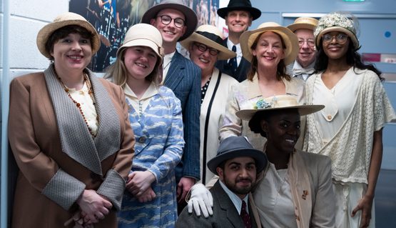 Backstage photo taken during Chariots of Fire. A group of performers wearing traditional 1920s clothing gather in a backstage corridor and smile.