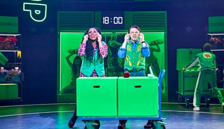 Georgina Onuorah (Audrey) and Colin Ryan (Seymour) in Little Shop of Horrors. Photo Manuel Harlan. Two people talk with a phone on each ear as they stand behind a trolley carrying large green boxes.