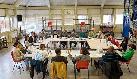 The Company of The Crucible in rehearsals. Photo by Manuel Harlan. Nineteen people are seated in a table arranged in a squared hoop. They chat and smile at each other.