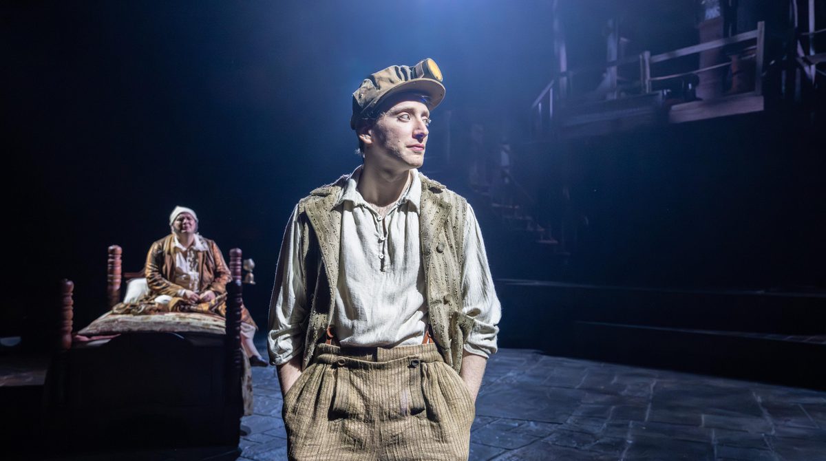 A person wearing beige trousers and waistcoat with a headlamp on their hat stands with their hands in their pockets. Behind, a person sits on a bed, looking upset.