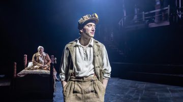 A person wearing beige trousers and waistcoat with a headlamp on their hat stands with their hands in their pockets. Behind, a person sits on a bed, looking upset.