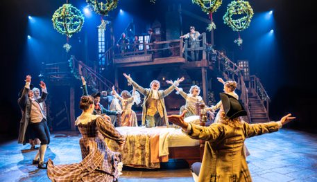 A raucous and joyful party of people dressed in bright Victorian clothing. Everyone sings and dances around a bed. Above, Mistletoe hangs in great baubles.