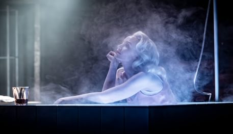 A person sits in a steaming bath and looks up thoughtfully, resting their hand on their chin. A shot of whiskey is on the edge of the bath