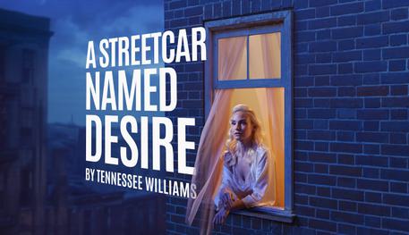 A dusky blue scene, focusing at a window high up on a building. A person with long blonde hair wearing a white silk dressing down leans on the windowsill and stares out with wonder. Next to the window, in white capital letters: A STREETCAR NAMED DESIRE BY TENNESSEE WILLIAMS