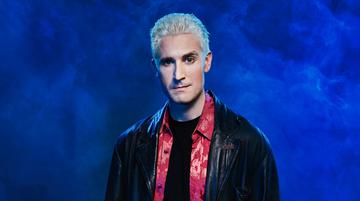 Brendan Murphy standing in front of a blue wall. He has bleached blonde hair and he is wearing a leather jacket.