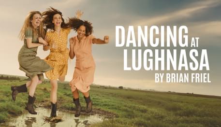 Three people with long hair and wearing pastel-coloured dresses smile and laugh as they hold hands and jump into a puddle in the middle of a grey green field. Text reads: DANCING AT LUGHNASA