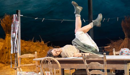 A person laughs as they lie across a kitchen table, both legs kicking in the air. They wear thick boots and a skirt that falls up as they move.