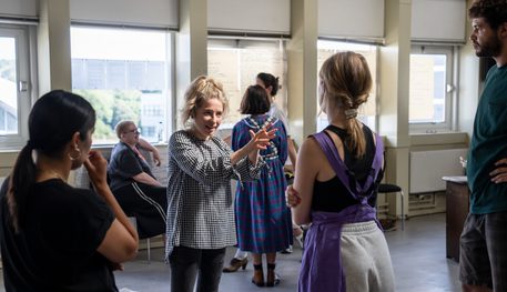 A person blonde hair wearing a tartan blouse holds both of their hands in front of them as they explain something to a group of people, who listen intently.