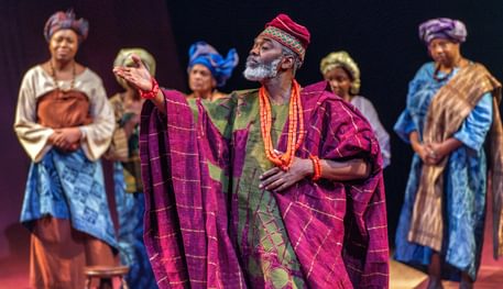 Wale Ojo and members of the company in Death and the King’s Horseman. Photo by Anthony Robling. A person closes their eyes and holds one hand up as if beckoning. They wear purple and green patterned clothing and orange jewellery. In the background, other people line up and watch them.