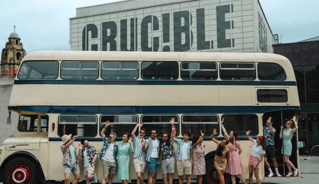 A large group of people wearing 1960s clothing smile and wave, standing in a line in front of a cream vintage double decker bus. Behind, the Crucible theatre.