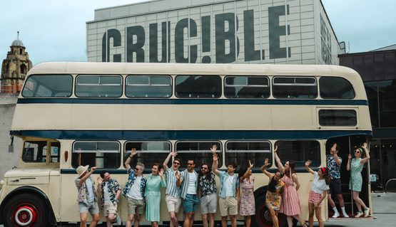 A large group of people wearing 1960s clothing smile and wave, standing in a line in front of a cream vintage double decker bus. Behind, the Crucible theatre.