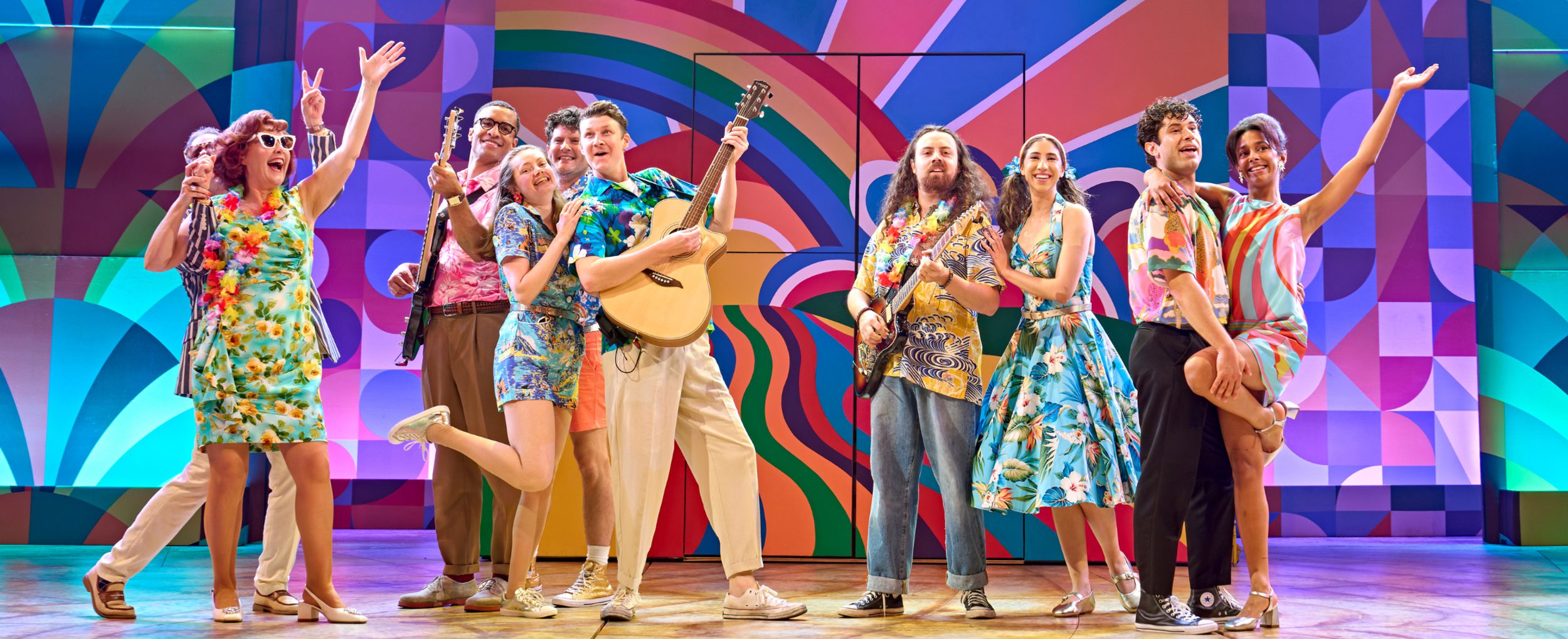 Members of the company in Summer Holiday the Musical. Photo by Manuel Harlan. A group of people wearing colourful, retro clothing smile, holding their arms in the air or playing musical instruments. Behind, a bright, technicolour set.