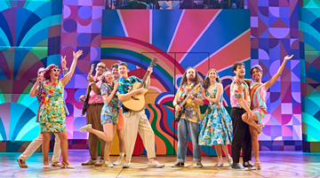 Members of the company in Summer Holiday the Musical. Photo by Manuel Harlan. A group of people wearing colourful, retro clothing smile, holding their arms in the air or playing musical instruments. Behind, a bright, technicolour set.