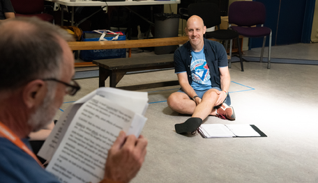 Robert Hastie (Director) in rehearsals for Much Ado About Nothing. Photo by Chris Saunders. Director Robert sits on the rehearsal room floor with an open script laid out in front of them. Robert smiles at an actor who is blurred in the foreground, reading from another script.