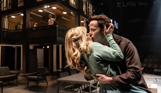 Rachael Wooding (Rose) and Robert Lonsdale (Harry) in Standing at the Sky’s Edge. Photo by Johan Persson. Rachael wears a blue 1950s dress and Robert wears a brown jacket. They kiss lovingly as they dance in front of a concrete structure behind them.