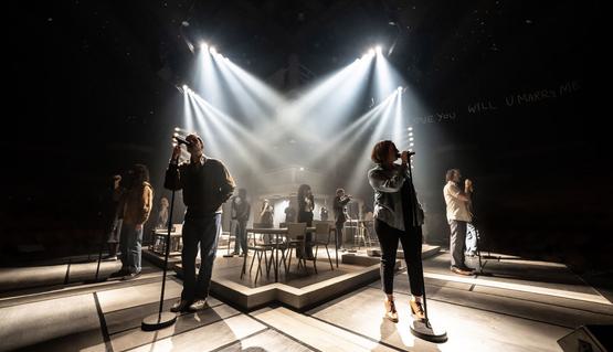The Company of Standing at the Sky’s Edge. Photo by Johan Persson. A full cast all stand with microphones on stands. They are dotted around the stage and are singing. They are back lit by bright lights shining down from the ceiling.