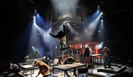 The Company of Standing at the Sky’s Edge. Photo by Johan Persson. A wide photo of a chaotic set. Lots of people are arguing, crying, fighting, shouting and holding their heads in their hands. A person leaps across a kitchen table with their arms in the air. Overlooking everything is a large concrete structure, shrouded in smoke and lights.