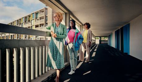 Three women stand on the Brutalist concrete balcony of Park Hill flats in Sheffield. A woman with blonde, backcombed hair wears a 1950s blue dress and gazes out at the view. Behind her, a young woman in 1980s jeans and jacket stares upwards. Behind her, a woman in a neutral formal suit also looks across. The sun shines and casts shadows across the balcony.