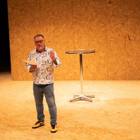 A man in a patterned shirt and jeans stands on stage with a script in hand, speaking outwards and gesturing with his free hand.