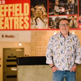 A man stands in the Crucible foyer, smiling at the camera with his hands in his jean pockets.