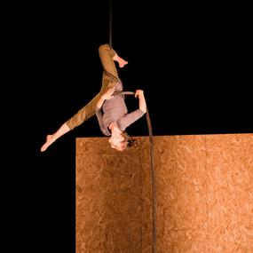 An aerial artist hangs from a rope from above. She is upside down, one leg wrapped around the rope, the other split, pointing toward the floor.