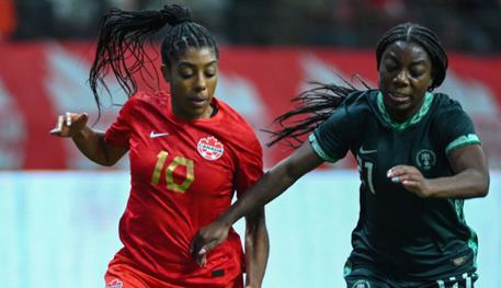 Two black women play football. One wears a red shirt and the other wears a black shirt. They are running across a football pitch.