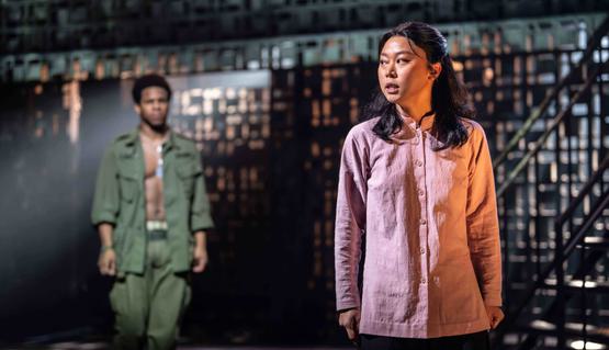 Jessica Lee (Kim and Alternate Mimi) and Christian Maynard (Chris) in Miss Saigon. Photo by Johan Persson. Jessica stands with hands by their side and looks slightly over their shoulder as they sing. Behind, Christian is blurred.