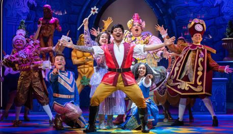 Duncan James (Danton) and the Company of Beauty and the Beast. Photo by Sam Taylor. Duncan holds their arms out widely and smiles. Behind, the rest of the company gather round and pose around him.