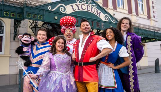 Promotional image for Panto 2023: the principal cast of Beauty and the Beast pose in front of the Lyceum entrance in panto costumes. Left to right are Max Fulham holding Gordon the Monkey, Jennie Dale as Fairy, Damian Williams as Madame Bellie Fillop, Duncan James as Danton, Bessy Ewa as Belle and Aidan Banyard as the Prince/Beast.