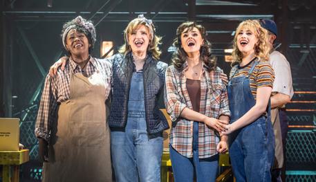 A group of women stand closely together, singing. They wear denim overalls and flannel shirts.