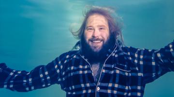 A person wearing a checked shacket, fully submerged underwater, smiles with their hands out to either side of them.