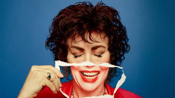 Promotional image of Ruby Wax. A close of Ruby holding a ripped up picture of the bottom half her face, which is smiling, over her mouth.