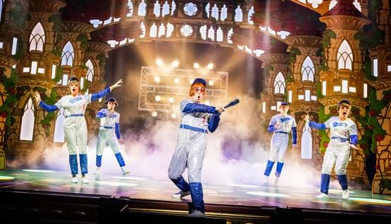 Marc Pickering and members of the company in Snow White Photo by Pamela Raith. A person in a sparkly baseball suit swings a bat and looks on in awe. Others dressed the same shout and a scream in joy