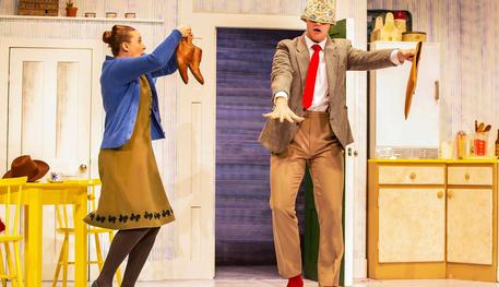 The Tiger Who Came To Tea production shot. Two actors performing centre stage. There is a woman on the left who is holding up shoes and a man who is trying to walk with an item over his head.